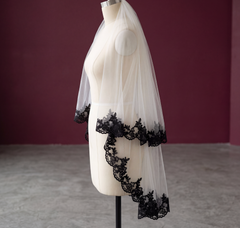 A white bridal veil with black lace trim draped over a cream dress form mannequin, set against a dark burgundy wall and gray floor.